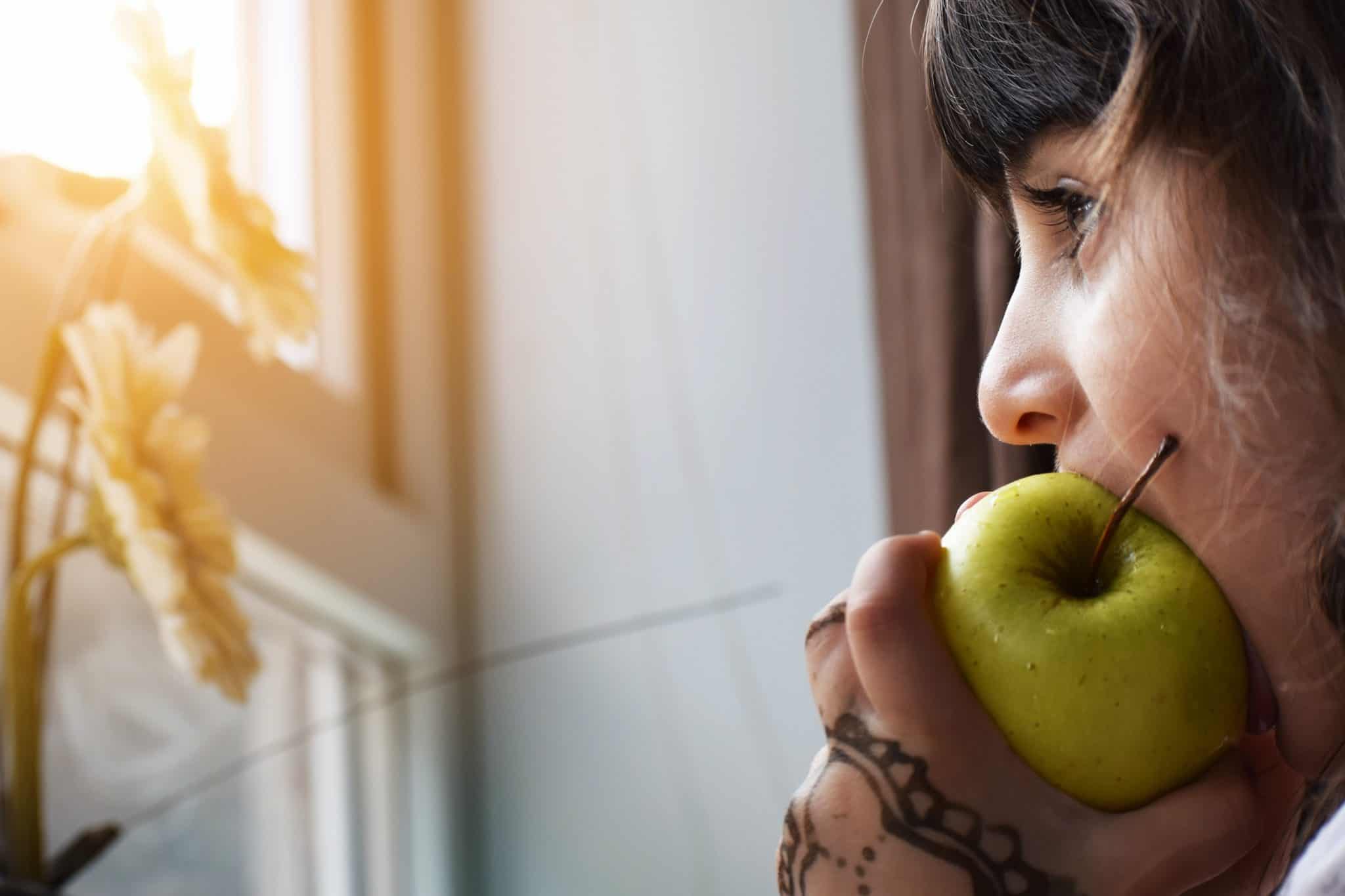 Girl with tattooed hand eating green apple by a window.