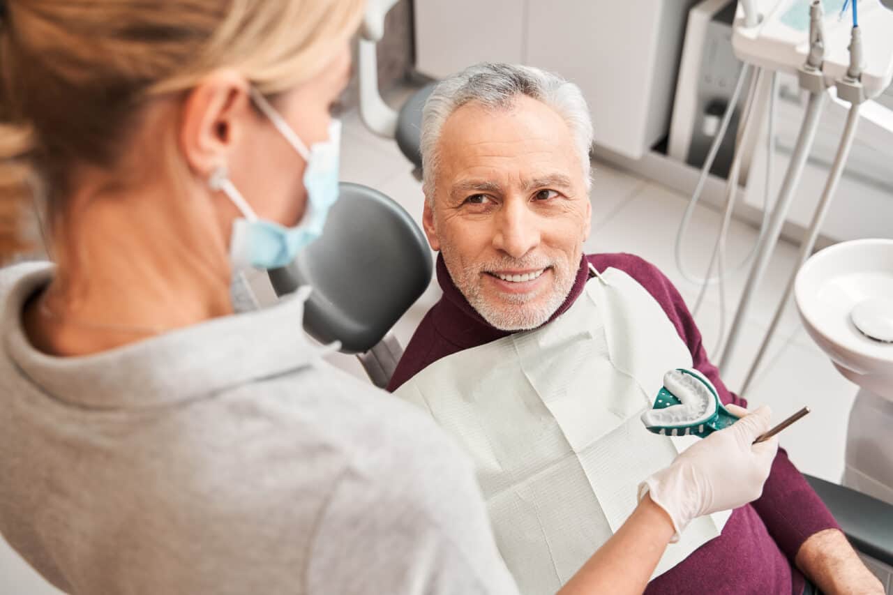 Back view of the female blonde caucasian doctor presenting dental impression for dentures to the senior smiling man. 
