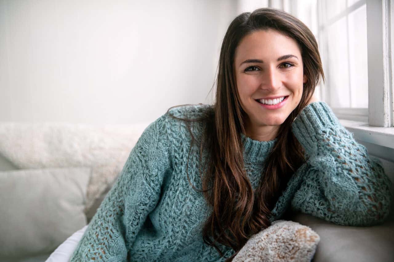 A young woman with long brown hair is sitting on a cozy couch near a window. She is smiling warmly and wearing a textured teal sweater. 