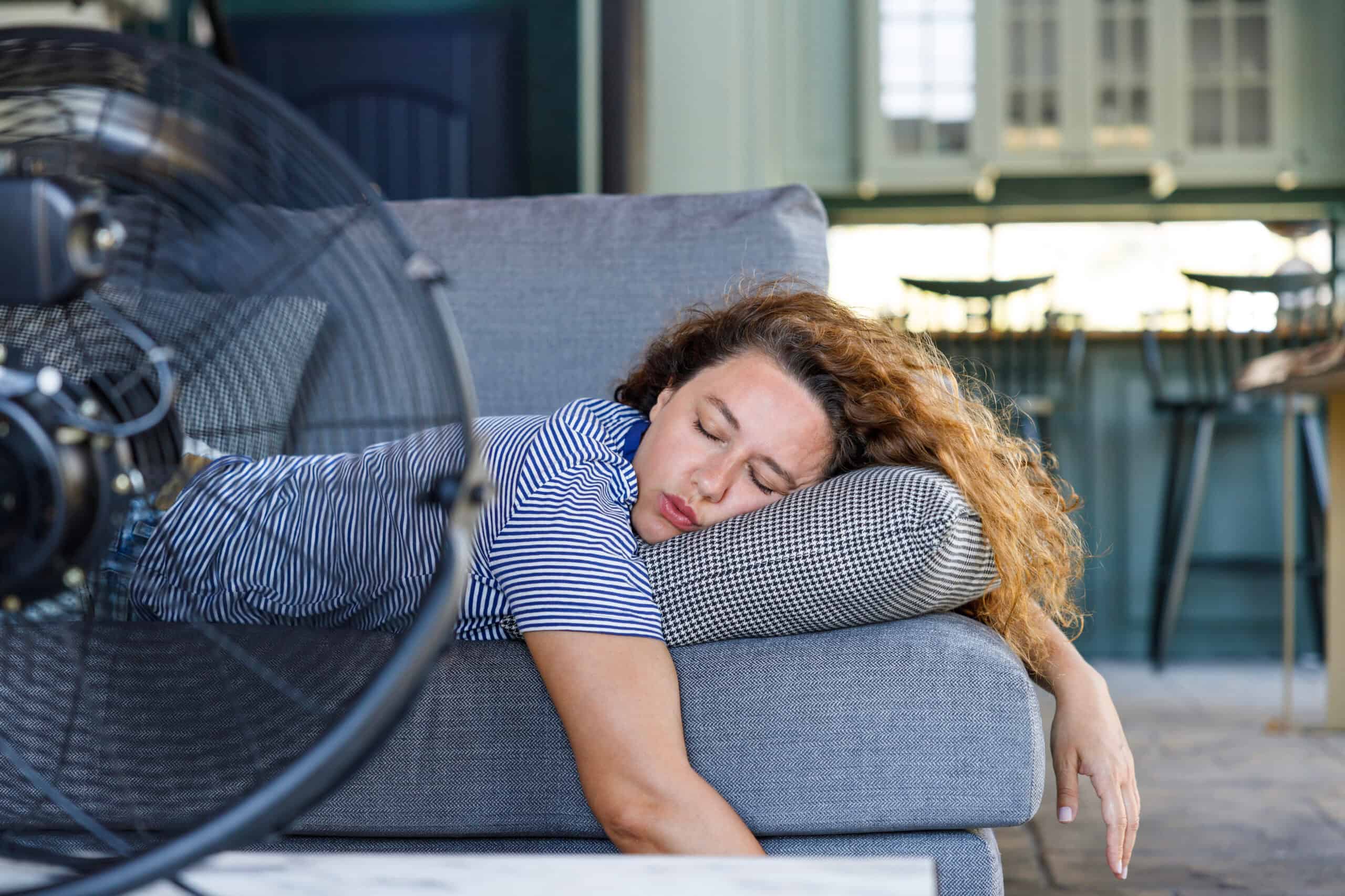 A woman laying in front of fan, hot and exhausted from the lack of sleep from the warmer environment.